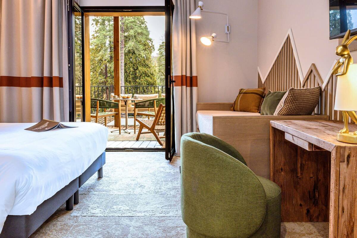 Ein Hotelzimmer mit Bett, Schreibtisch, Stuhl, einem Balkon mit Tisch und Stühlen sowie Blick auf Bäume.