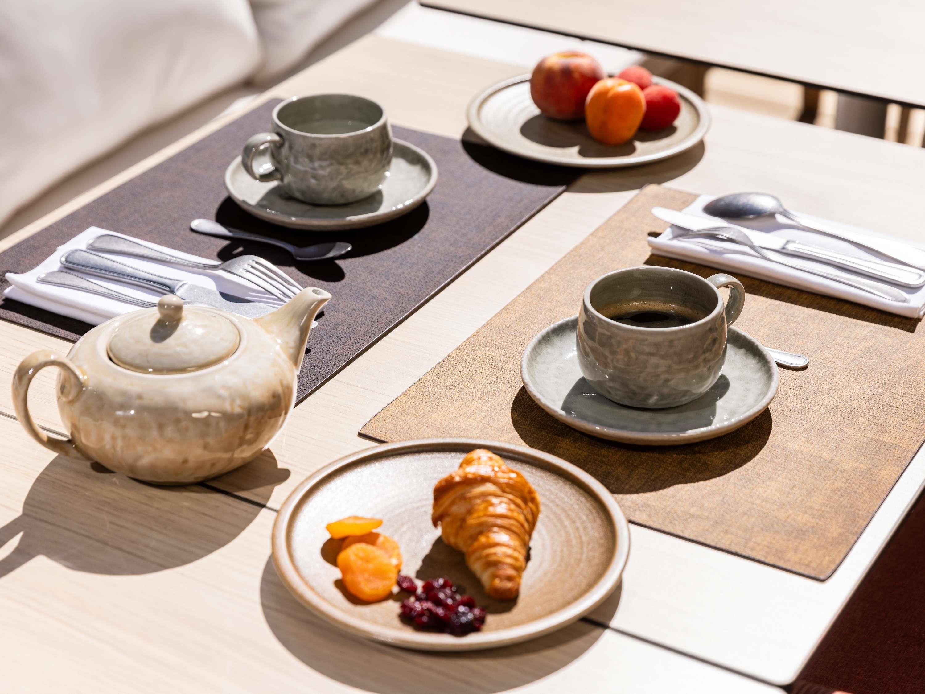 A breakfast setup with a teapot, cups, plates with fruits, and croissants on a table.