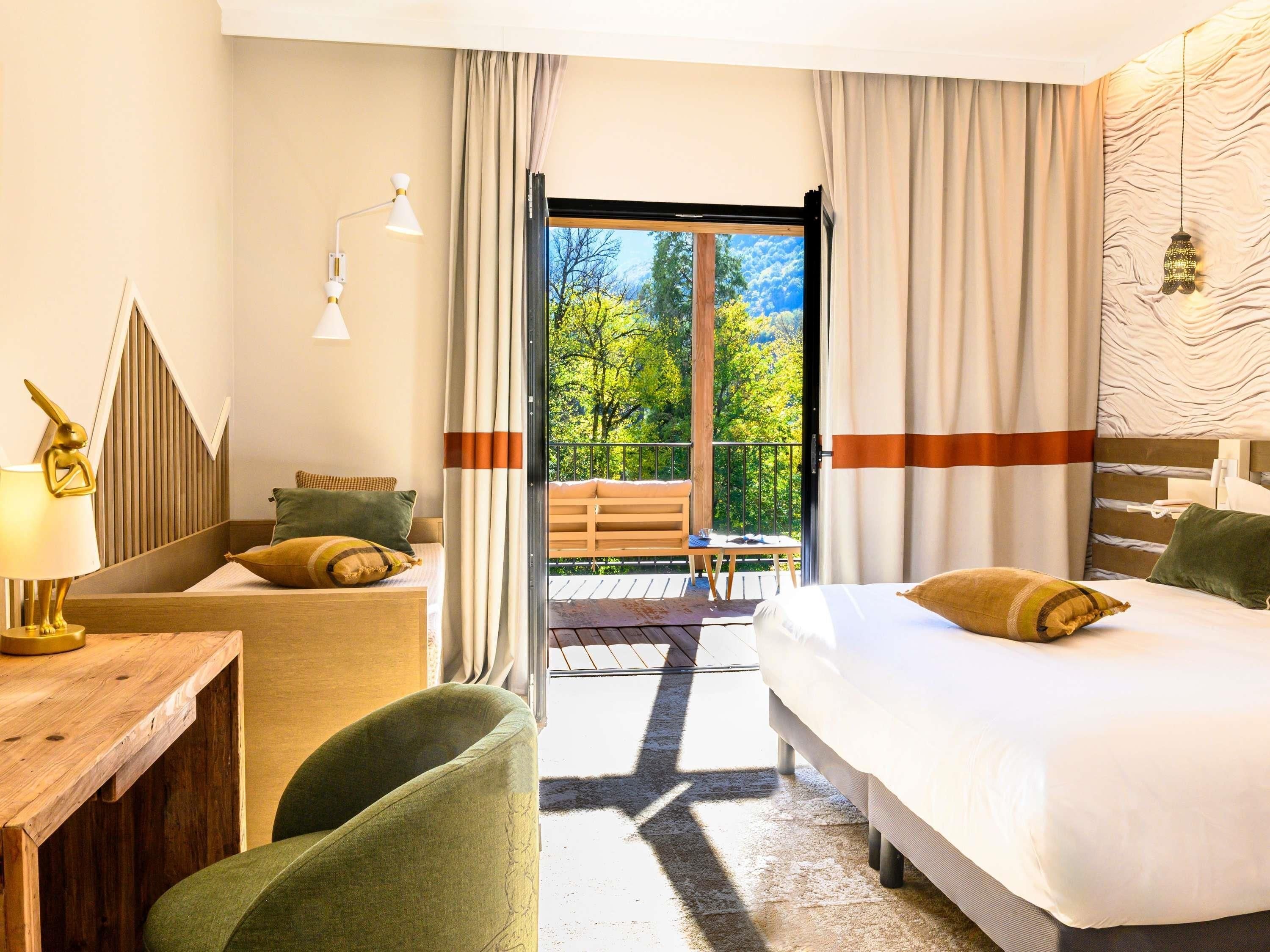 A hotel room with a bed, a desk, a chair, and a balcony with a view of trees.