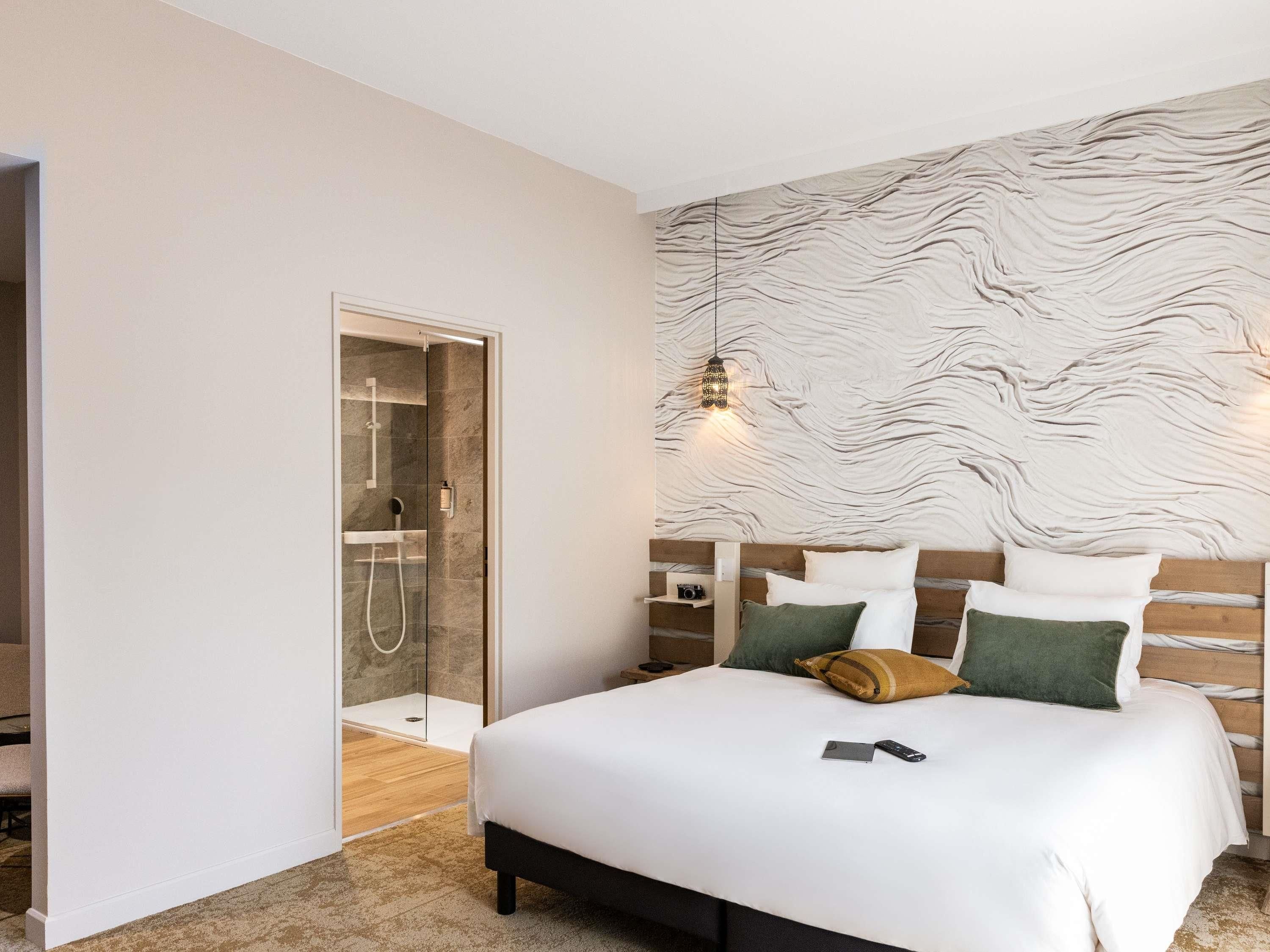 A modern hotel room with a large bed, a wooden headboard, and a glass-enclosed shower.