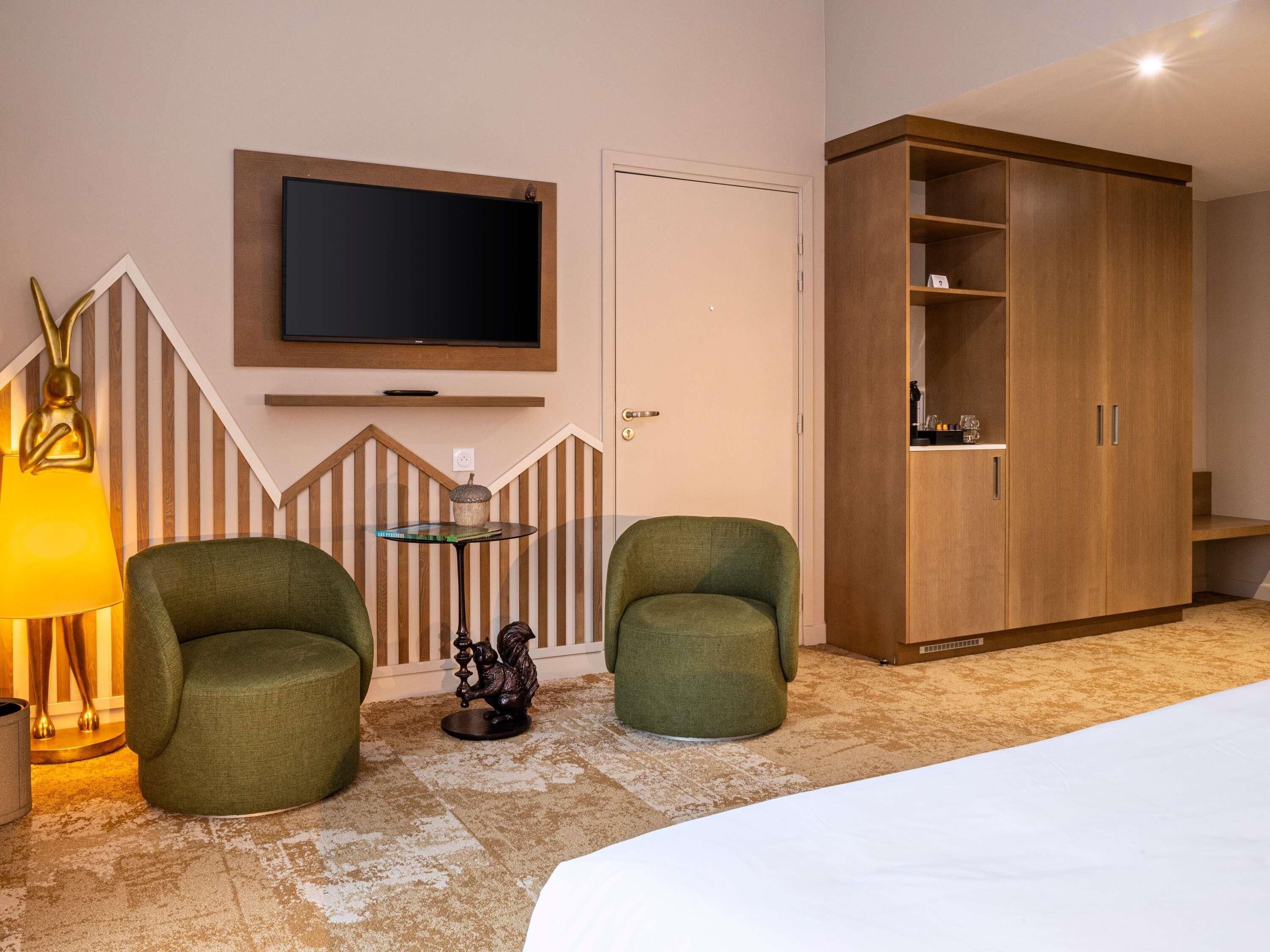 Een hotelkamer met een bed, twee groene fauteuils, een klein rond tafeltje met een boek, een wandgemonteerde televisie en een houten garderobe.