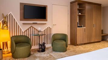 Een hotelkamer met een bed, twee groene fauteuils, een klein rond tafeltje met een boek, een wandgemonteerde televisie en een houten garderobe.