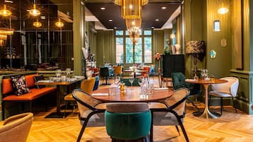 Een restaurant met een centraal eetgedeelte met ronde tafels en stoelen, een bar met krukjes, en een uitzicht op het buitenlands gebied door grote ramen.