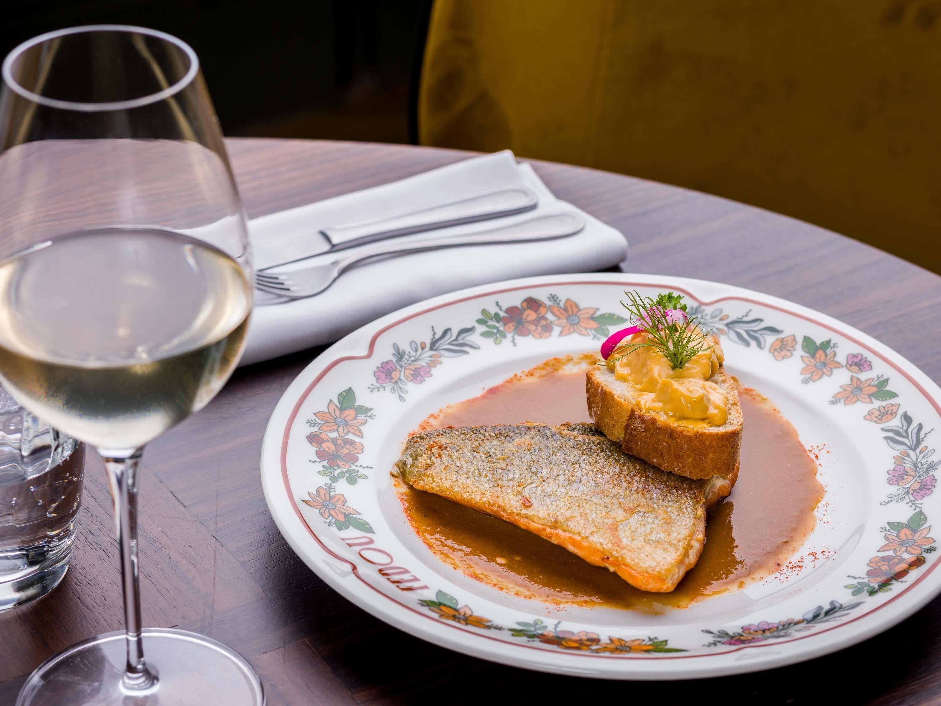 Un plato con un plato de pescado y salsa, una copa de vino blanco y una servilleta con cubiertos sobre una mesa de madera.