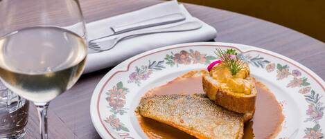 Un plato con un plato de pescado y salsa, una copa de vino blanco y una servilleta con cubiertos sobre una mesa de madera.