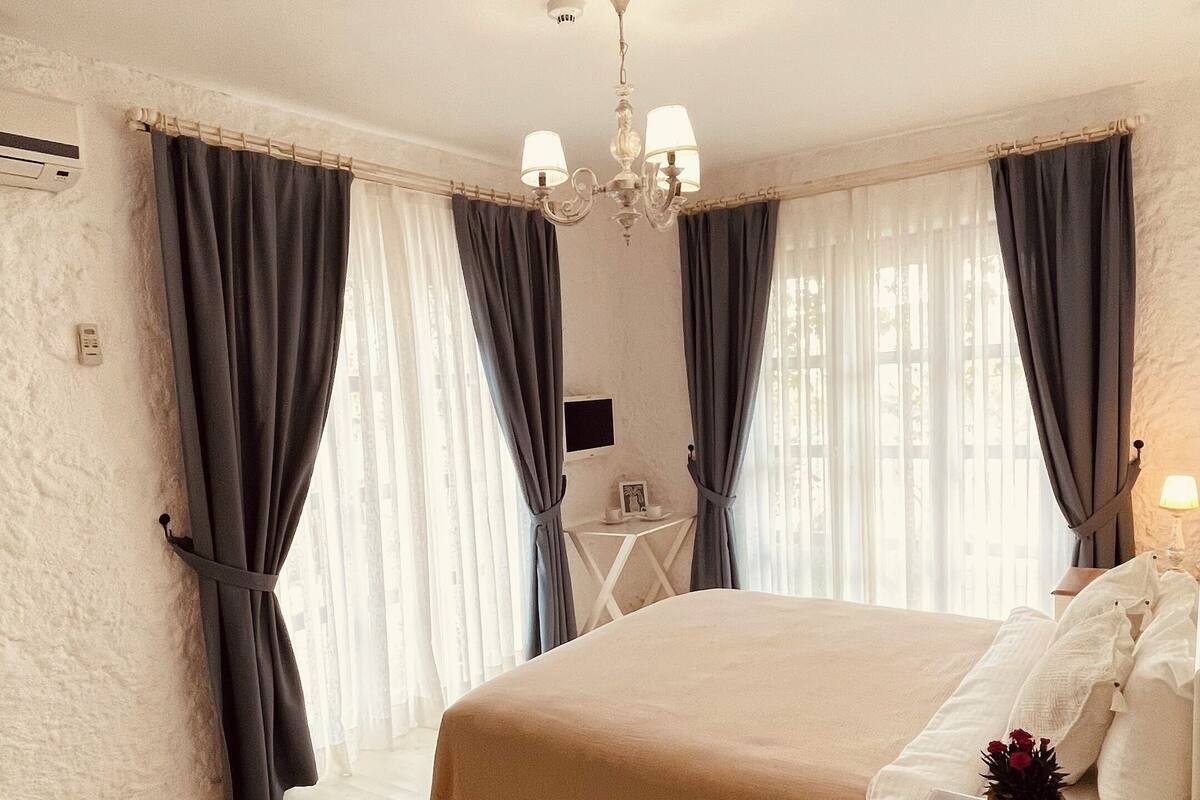 Un dormitorio con cama, una mesita de noche con lámpara, una pequeña mesa con un marco de fotos y una planta en maceta.