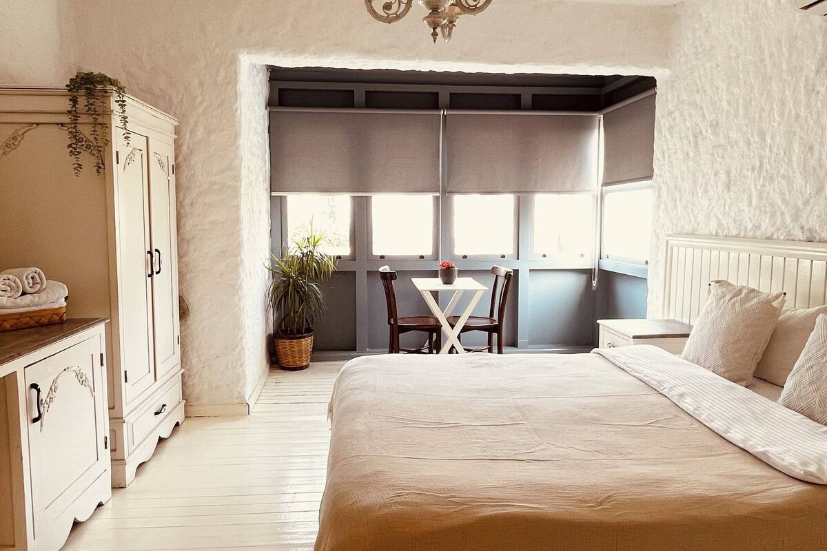 Un dormitorio con una cama grande, un candelabro, una pequeña mesa con sillas y vistas a través de una ventana.
