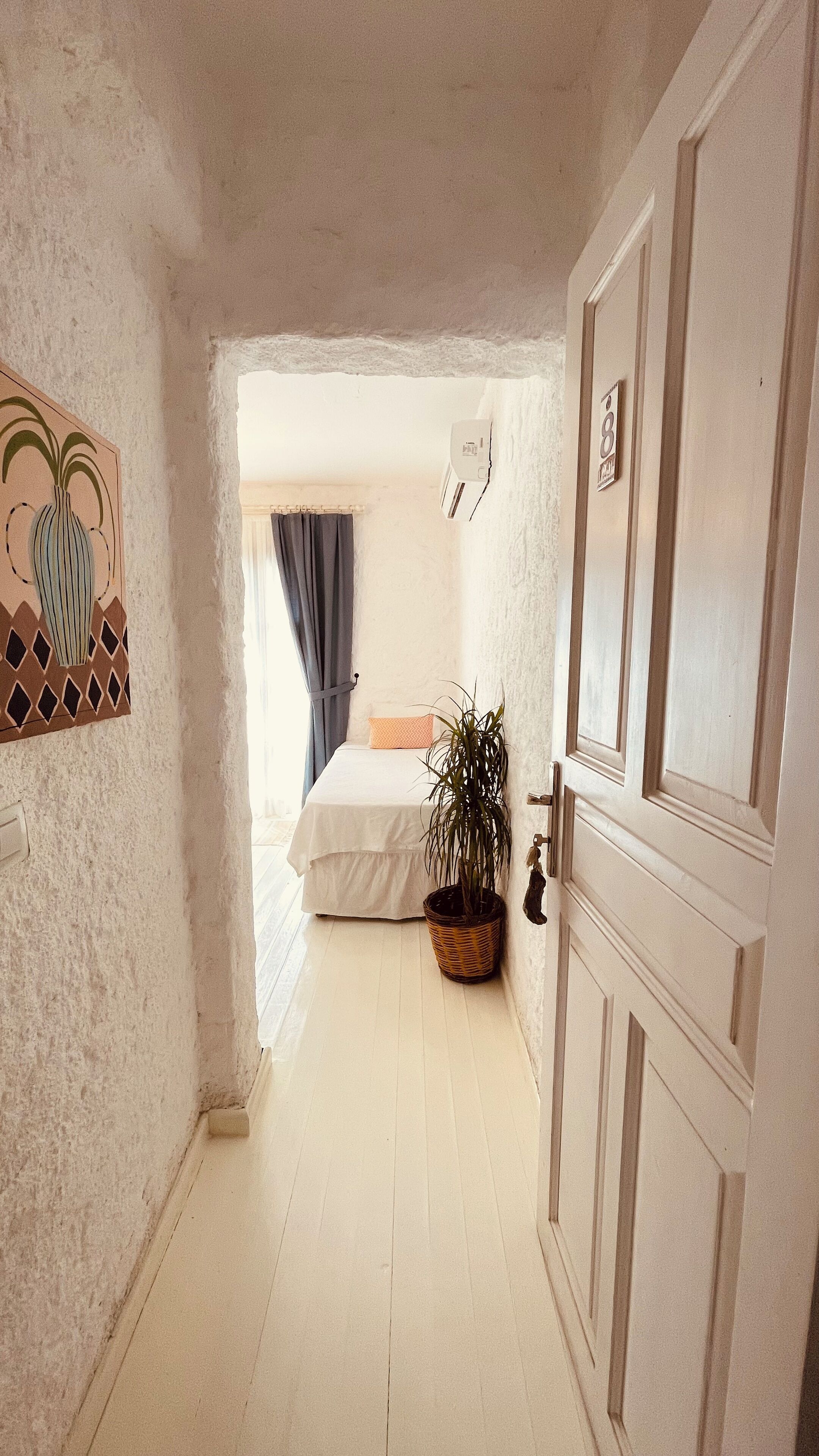 Ein schmaler Flur, der zu einem Schlafzimmer führt, mit einem Bett, einer Zimmerpflanze und einer Tür mit einer Nummer.'