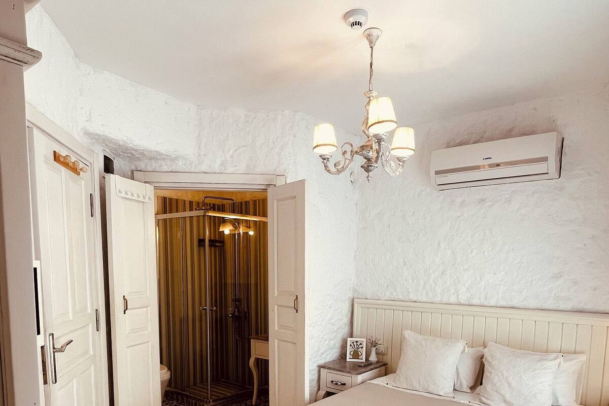 Un dormitorio con una cama grande, una mesita de noche con lámpara, ventilador de techo y un baño visible a través de una puerta abierta.