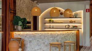 Zona de recepción con pared de piedra, techo de madera y mostrador de recepción. Hay dos sillones de mimbre y un jarrón grande.