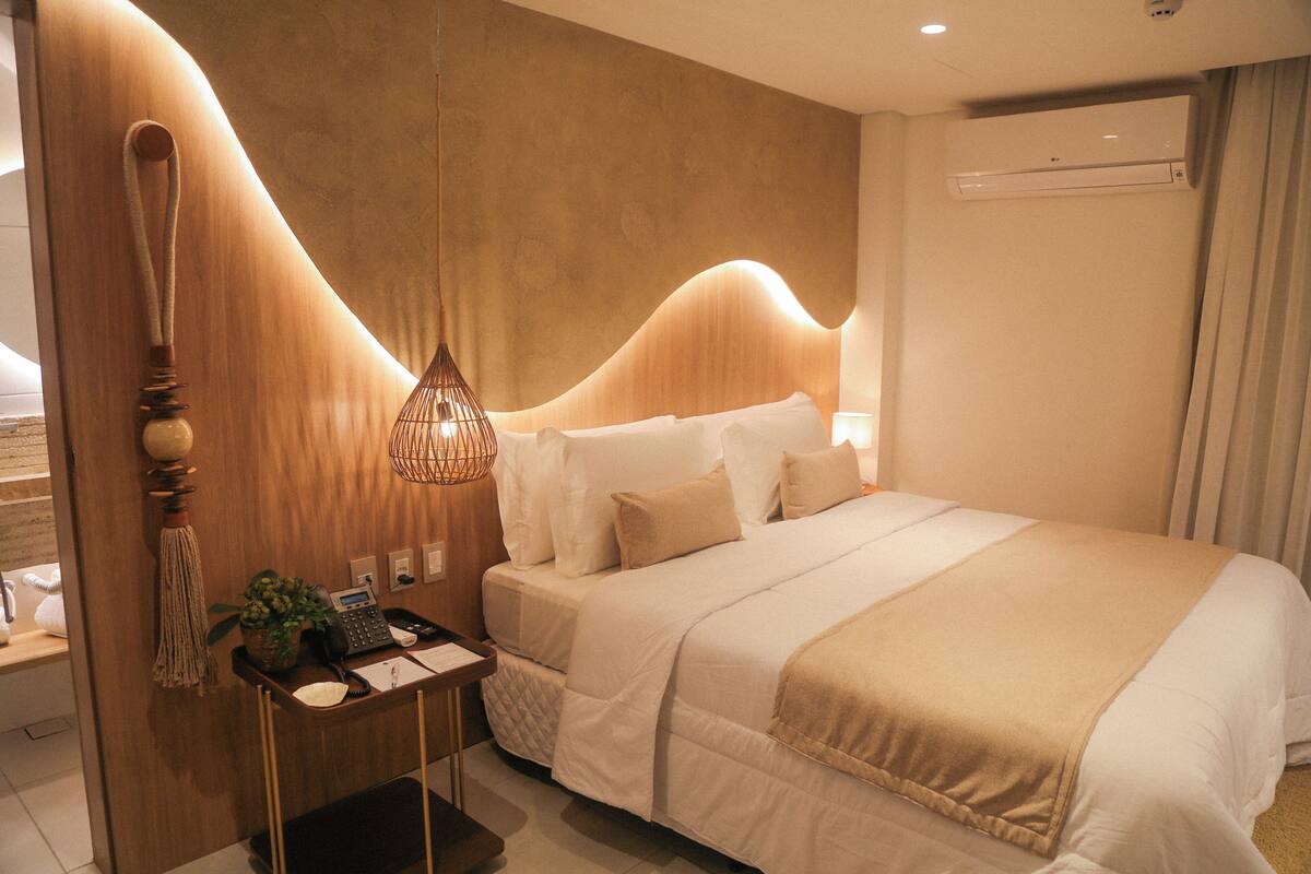 Quarto de hotel moderno, com uma cama grande, uma mesinha de cabeceira com abajur e ar condicionado fixado na parede.