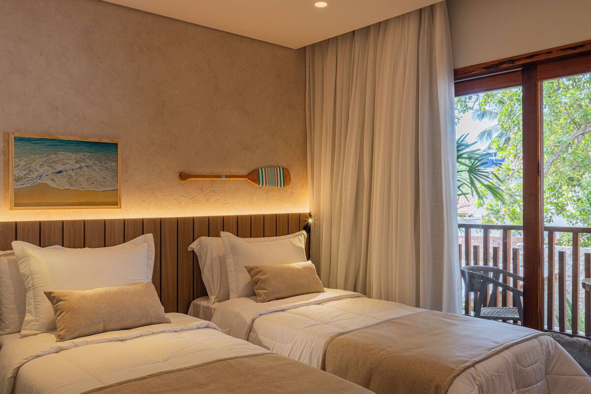 Quarto de hotel com duas camas, cabeceira de madeira, pintura de uma praia e vista para uma varanda com cadeira.