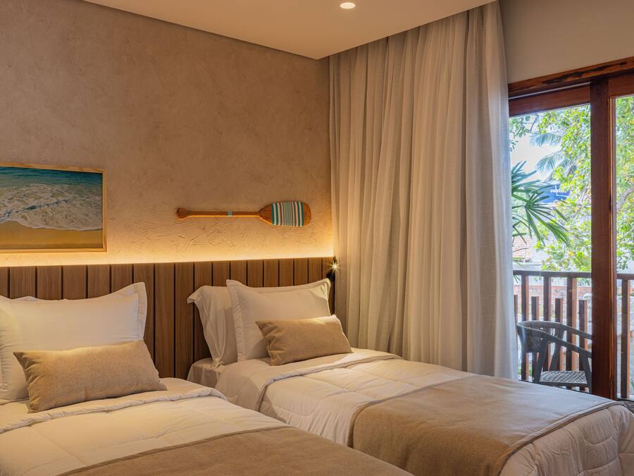 A hotel room with two beds, a wooden headboard, a painting of a beach, and a view of a balcony with a chair.