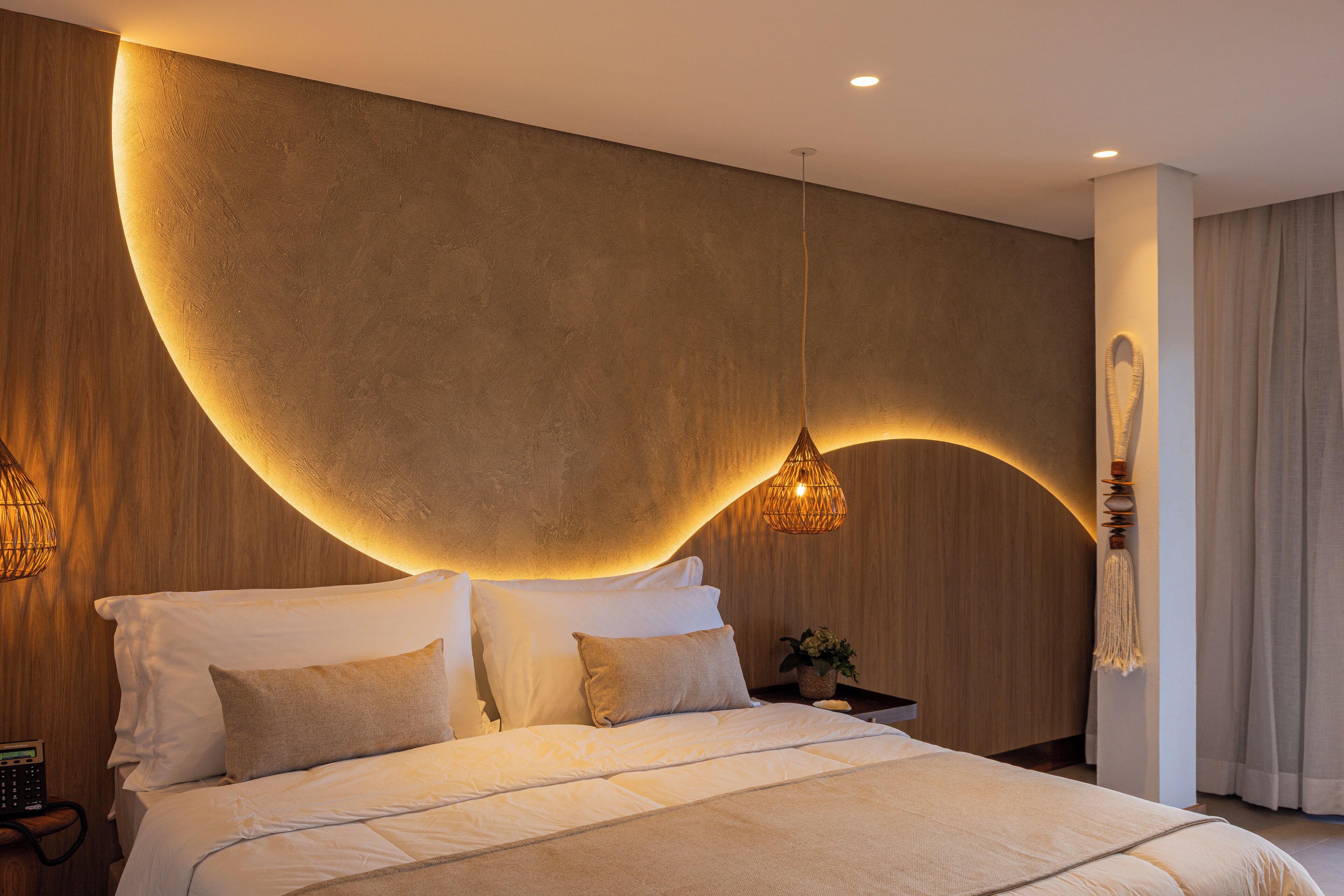 Un dormitorio moderno con diseño de pared curva, una cama grande con ropa blanca, dos lámparas de noche y una planta pequeña en la mesita.