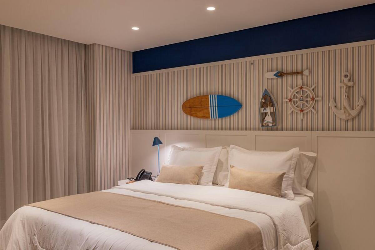 Quarto de hotel com uma cama grande, cortinas listradas e decoração náutica, incluindo uma prancha de surf, leme de navio e ancora.