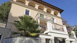 A two-story building with a balcony, a stone wall, and a palm tree in the foreground. - Your Breathtaking Penthouse (Gargnano)