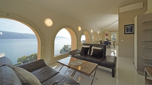 A modern living room with a large arched window overlooking a lake, a leather sofa, a glass coffee table, and a dining area in the background. - Your Breathtaking Penthouse (Gargnano)