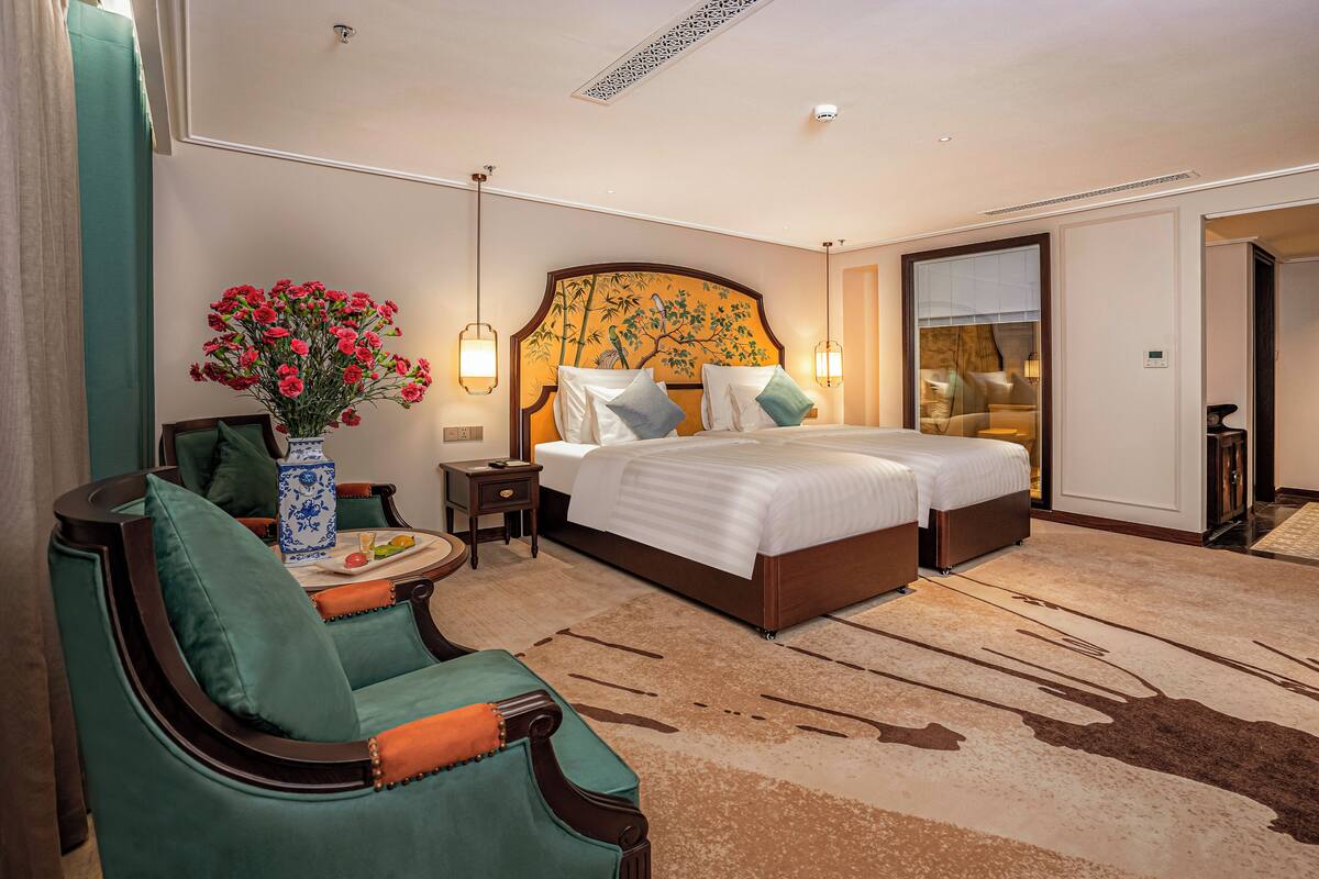 A hotel room with a large bed, two bedside tables, a chair, a vase of flowers, and a view of the outside.