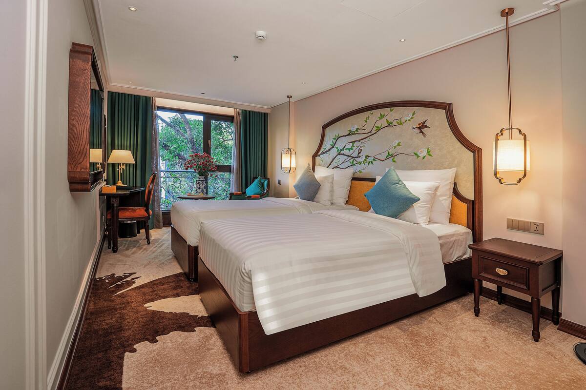 A hotel room with two beds, a nightstand, a lamp, and a view of greenery outside.