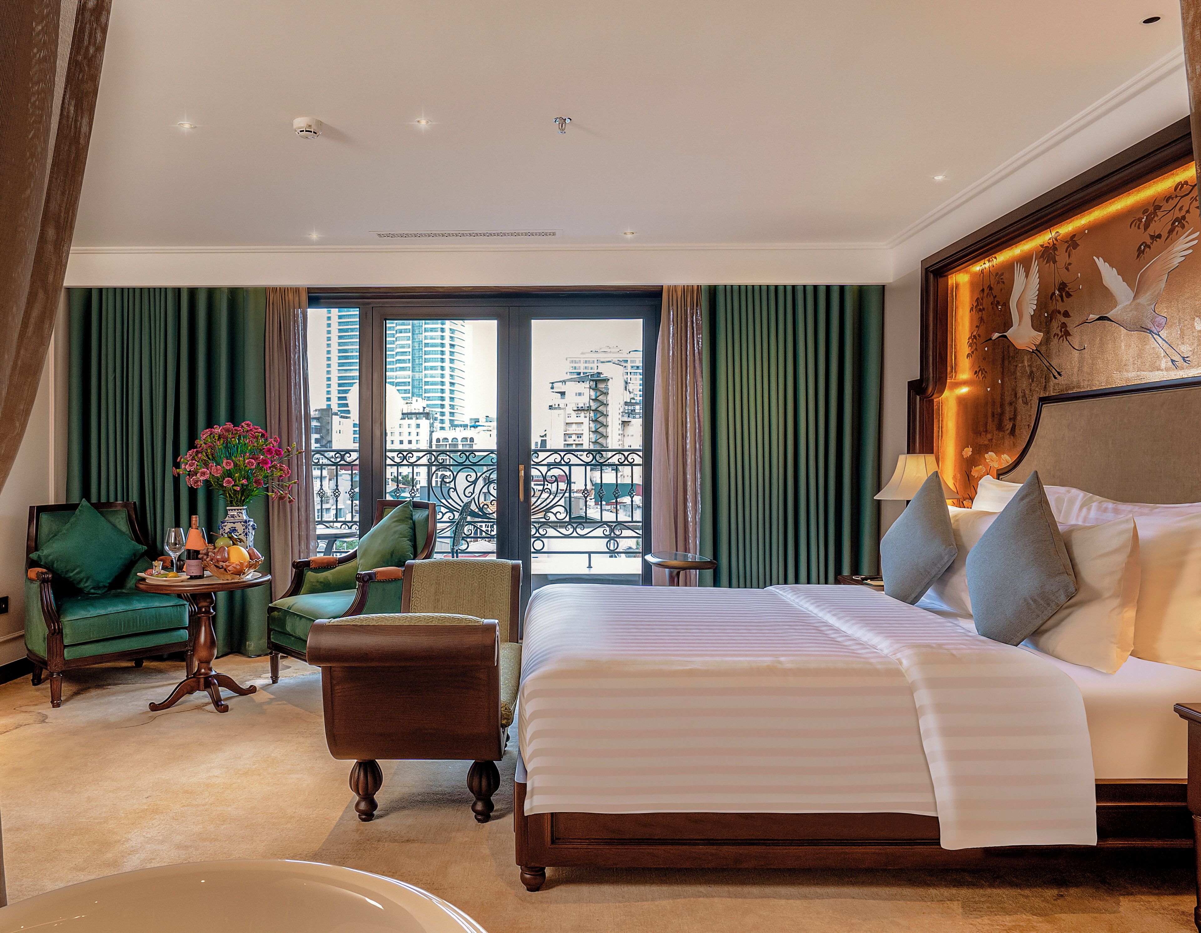 Una camera d'albergo con un letto grande, un angolo salotto con una poltrona e un tavolino, vista sulla città attraverso ampie finestre e un balcone con ringhiera.