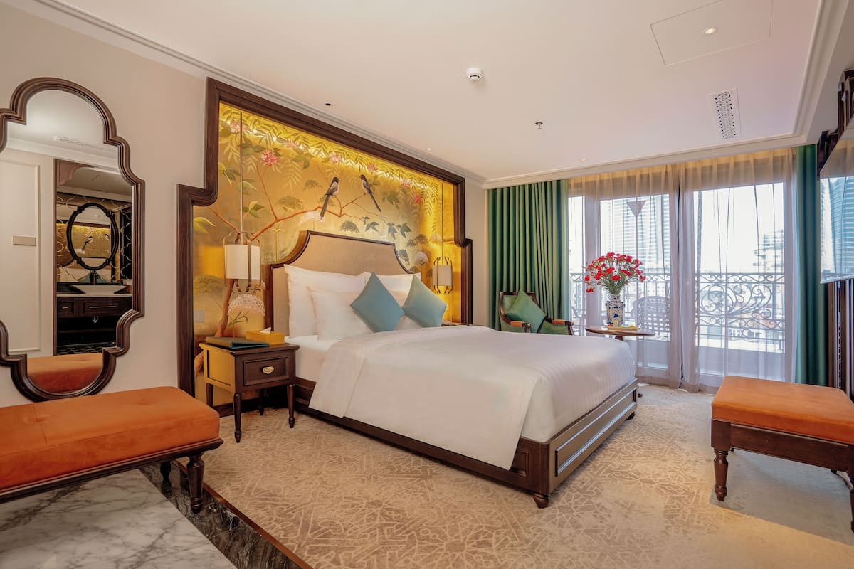 A hotel room with a large bed, a bedside table, a bench, a mirror, and a view of the city through large windows.
