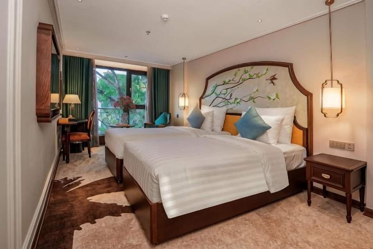 A hotel room with a large bed, bedside tables, a desk with a chair, and a view of greenery outside.