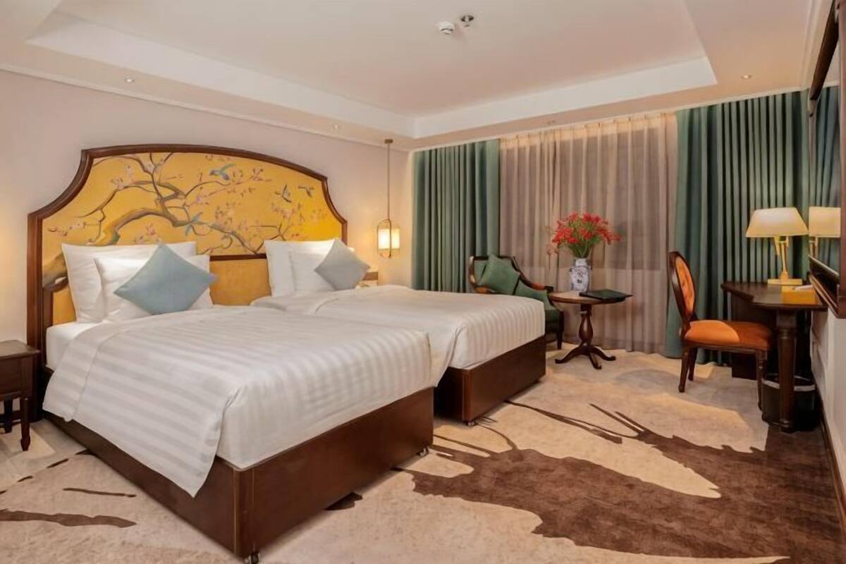 A hotel room with a double bed, a small table with a lamp, a chair, and a decorative vase with flowers.