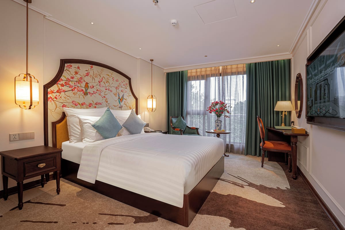 A hotel room with a large bed, a nightstand, a desk with a chair, a lamp, and a view of the city through the window.