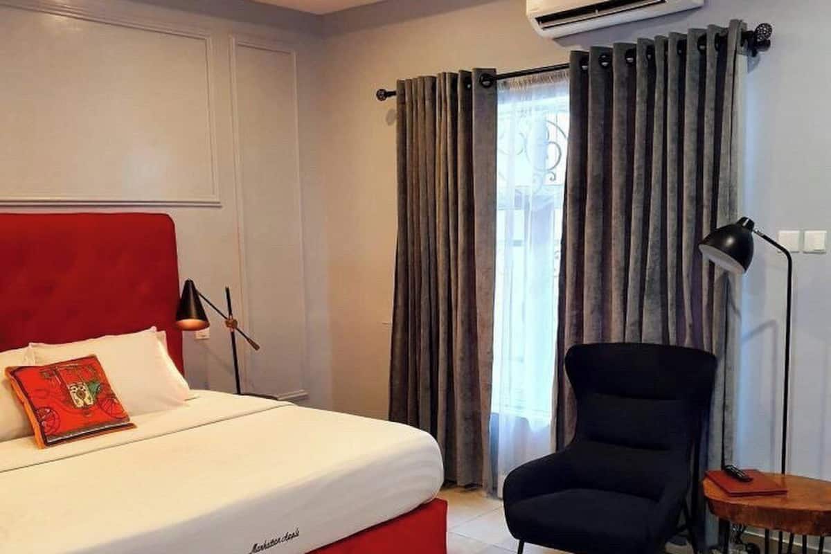 A hotel room with a red bed, a black chair, a small table, and a window with curtains.