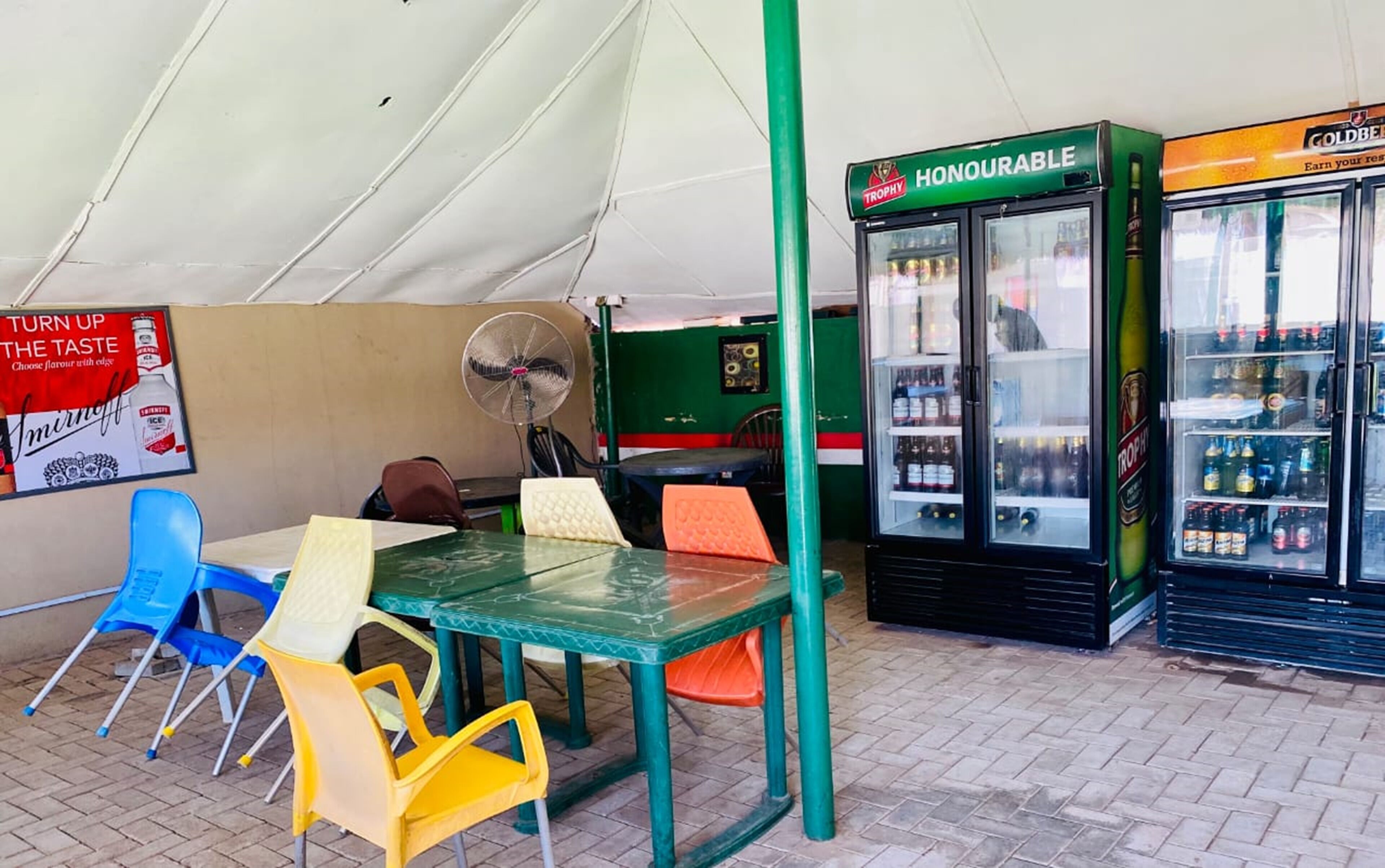 An outdoor seating area with a table and chairs, a fan, and a refrigerator stocked with beverages.