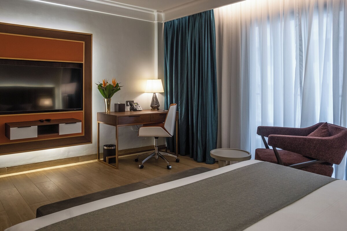 A hotel room with a large bed, a desk with a chair, a television, a lamp, and a vase with flowers.
