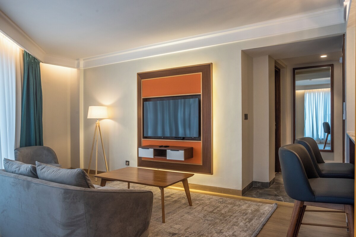 A modern hotel room with a flat-screen TV mounted on the wall, a sofa, a coffee table, and a seating area with chairs.