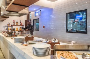A buffet setup with various food items and condiments, a chef in uniform, and a large window reflecting the kitchen area. - Okash City Boutique Hotel (Nairobi)