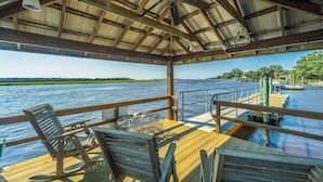 Une terrasse en bois avec une table et des chaises, donnant sur une étendue d’eau dont on aperçoit la rive au loin.
