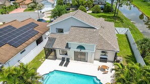 Ein zweistöckiges Haus mit Swimmingpool, Solaranlage und einem von einem Zaun umgebenen Garten mit Palmen.