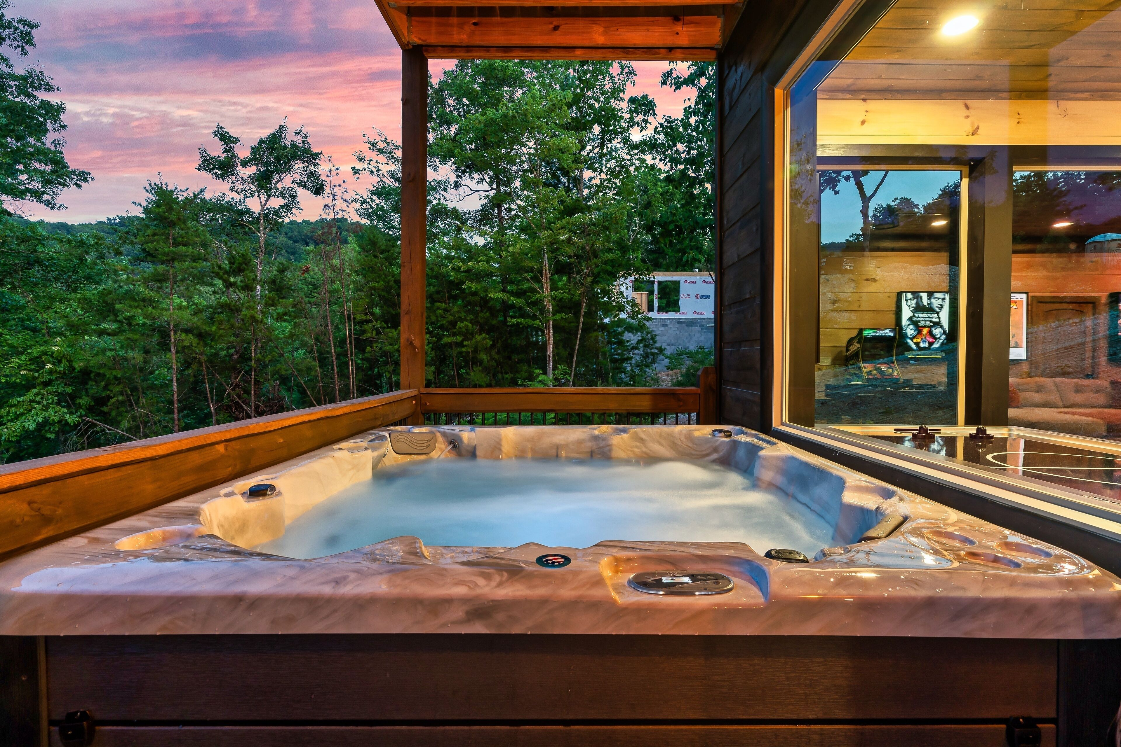 Ein Whirlpool mit Holzterrasse und Glaswänden, umgeben von Bäumen und Blick auf einen Sonnenuntergang.