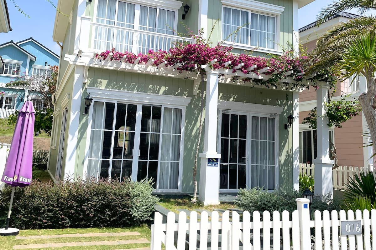 A two-story house with a white picket fence, a porch with pink flowers, and a clear sky.