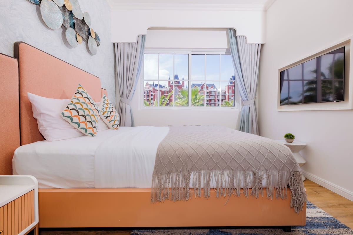 A bedroom with a bed, a window with a view of buildings, a TV on the wall, and a small plant on the nightstand.