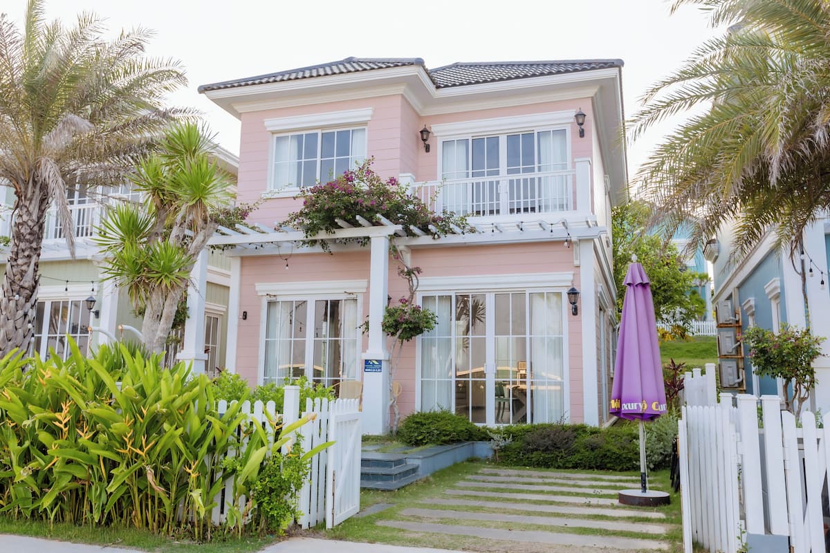 A pink house with a white porch, a small garden, and a purple umbrella.