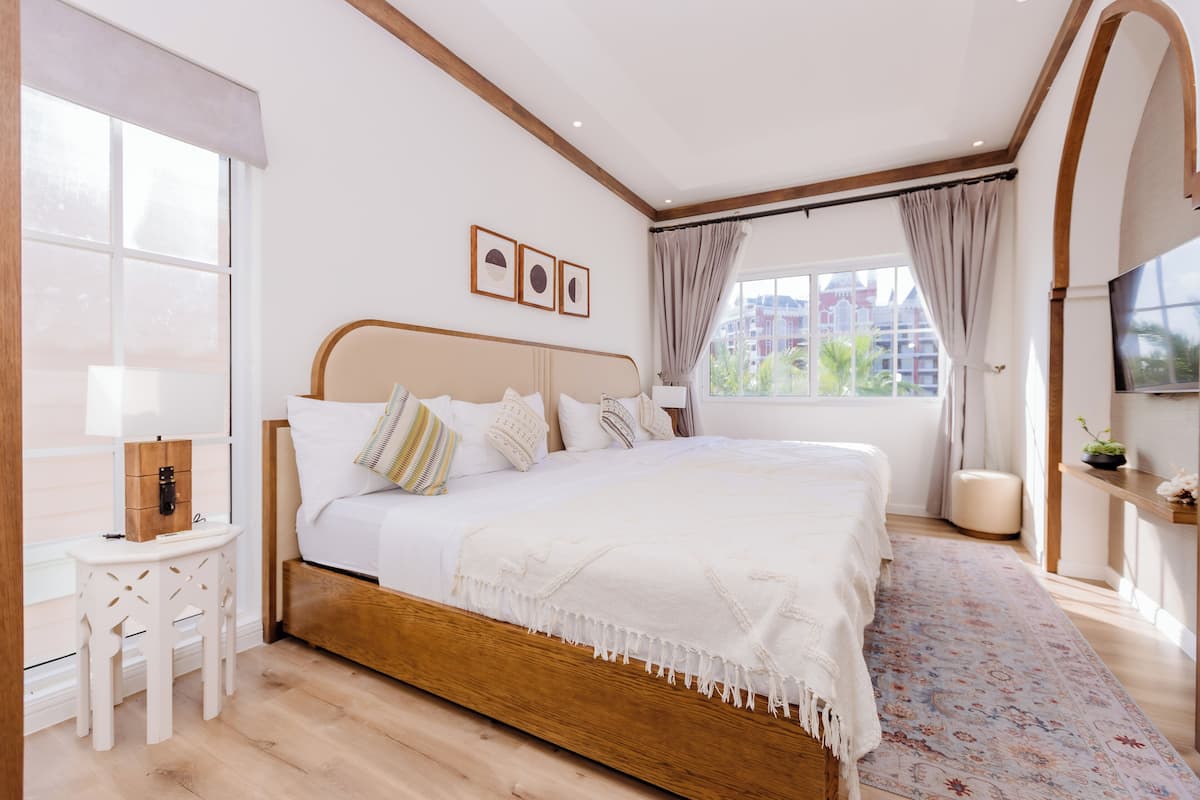 A bedroom with a wooden bed, white bedding, a small table with a lamp, and a view of buildings through large windows.