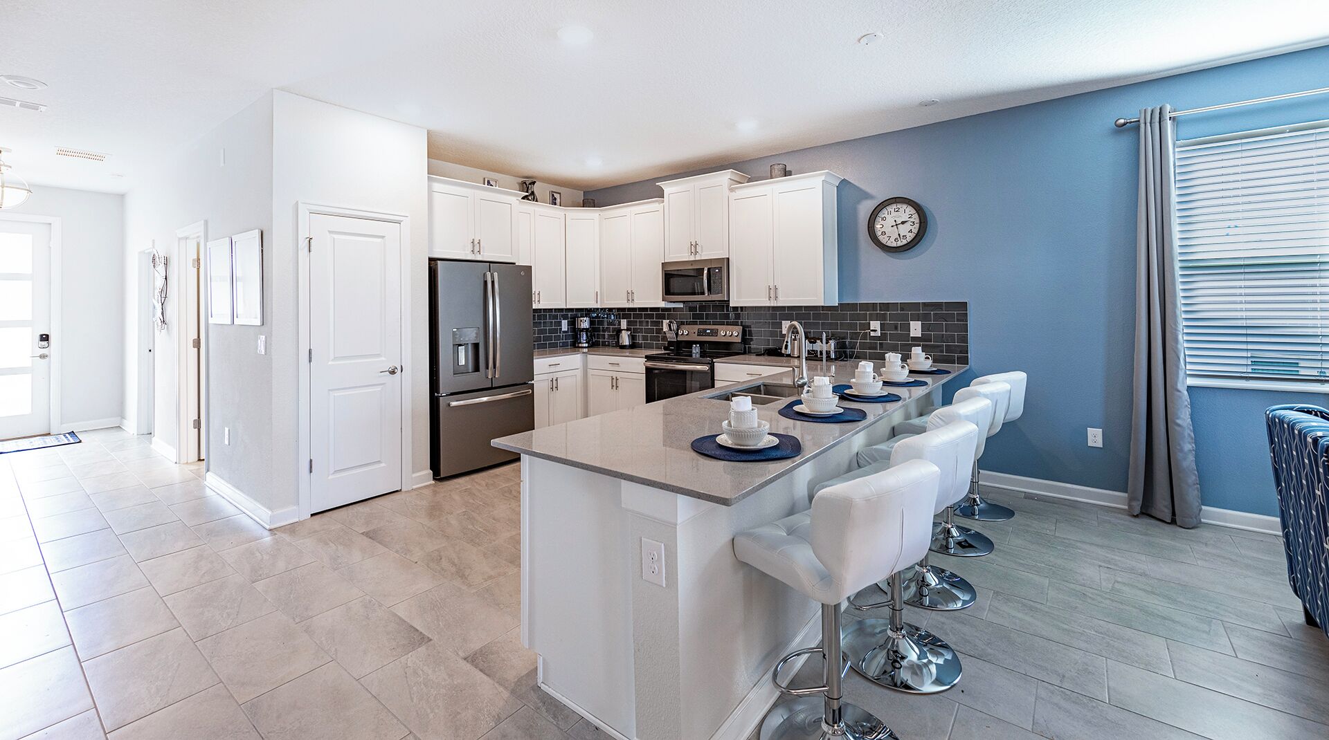 A modern kitchen with white cabinetry, a stainless steel refrigerator, a microwave, and a coffee maker. There is a breakfast bar with white bar stools and a clock on the wall.