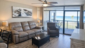 A living room with a sofa, armchair, and coffee table, a ceiling fan, and a large window with a view of the ocean. - Aransas Bay Condo with Balcony, Pool & Lighted Pier (Rockport)