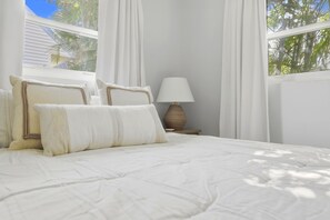 Ein Bett mit weißer Bettwäsche und zwei Kissen, eine Nachttischlampe und Blick auf Palmen durch ein Fenster mit weißen Vorhängen.