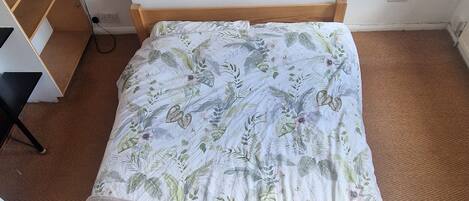 Una cama individual con una colcha estampada y una manta beige, cabecero de madera y suelo cubierto de alfombra marrón.