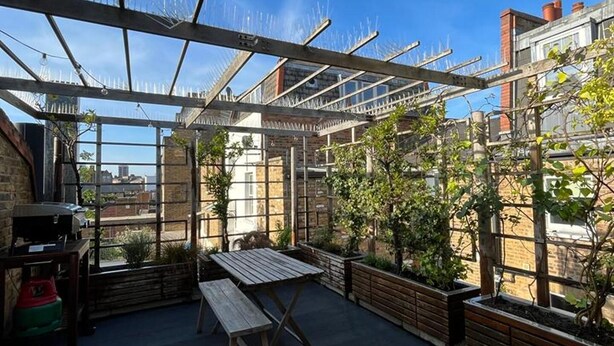 A rooftop patio with a wooden table and benches, surrounded by planters and a metal pergola structure.
