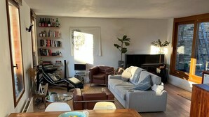 A cozy living room with a wooden dining table, two chairs, a grey sofa, a television, and a bookshelf filled with books and bottles.