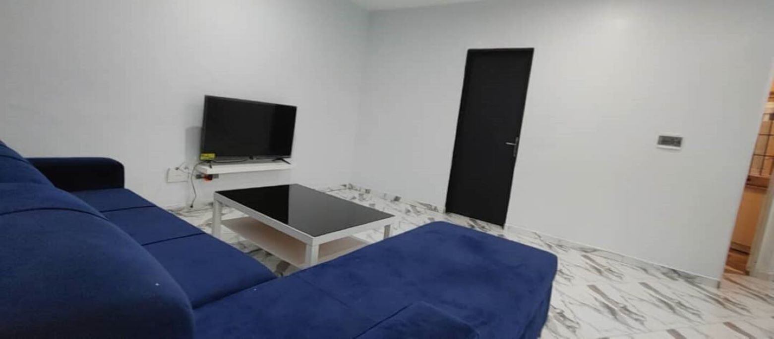 A room with a blue sofa, a small table, a TV mounted on the wall, and a door.