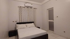 A bedroom with a bed, a nightstand, an air conditioning unit, a patterned curtain, and a door. - 2 Bedroom Furnished Living Room, Air-conditioned Legbassito Lomé


 (Lome)