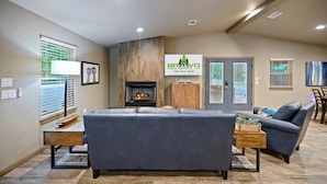 A modern living room with a fireplace, a sofa, a dining area, and a sign for Bravo Cabin Rentals, LLC.