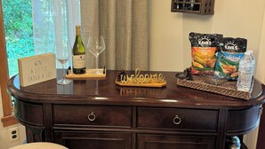 Zona de bar con una botella de vino, copas, un letrero de bienvenida, aperitivos y un estante pequeño con utensilios de cocina.